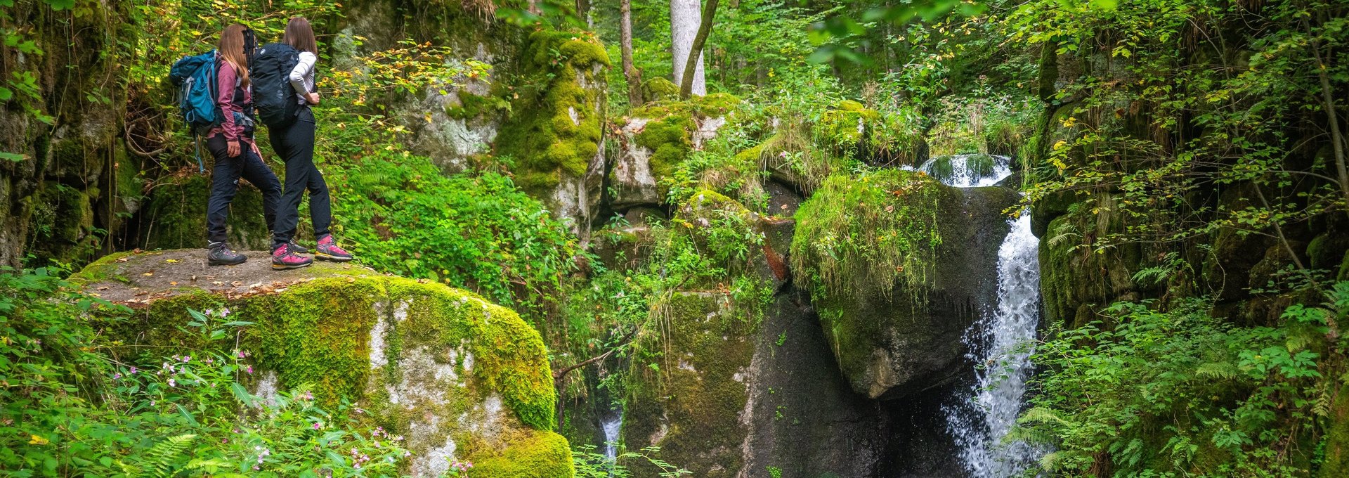Zwei Wanderer stehen auf einem Fels vor einem Wasserfall